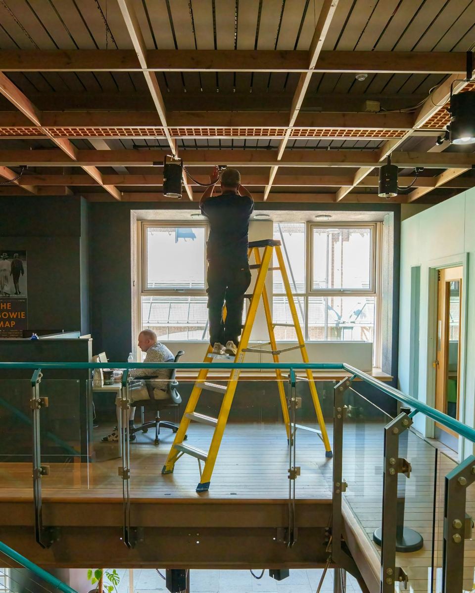 Engineer on a ladder wiring a fire alarm loop at ceiling height in an open-plan office